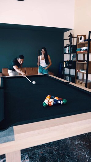 A pool table with balls, a person playing, and a bookshelf in a room.