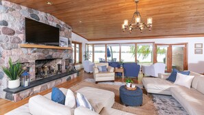 A spacious living room with a stone fireplace, a large flat-screen TV, and a wooden ceiling. There are multiple seating areas with sofas and chairs, and large windows offering views of the outdoors.