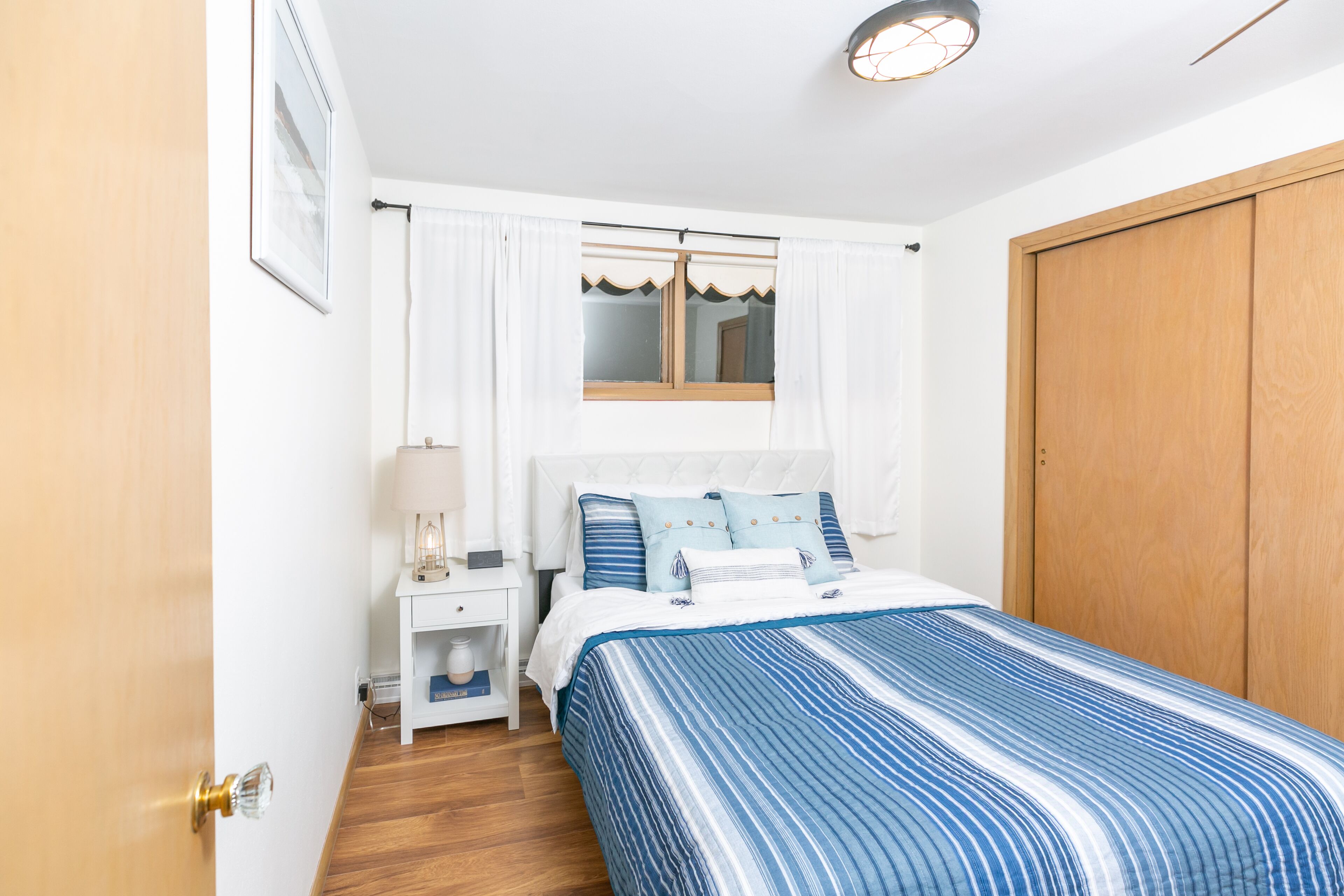 A bedroom with a bed, a nightstand, a lamp, a window with curtains, and a wooden wardrobe.