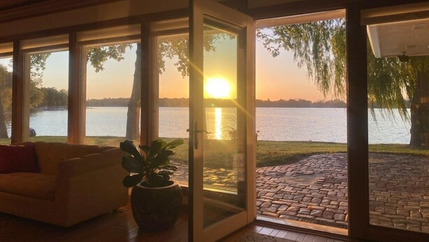 A room with a large sliding glass door leading to a patio with a view of a lake and sunset.