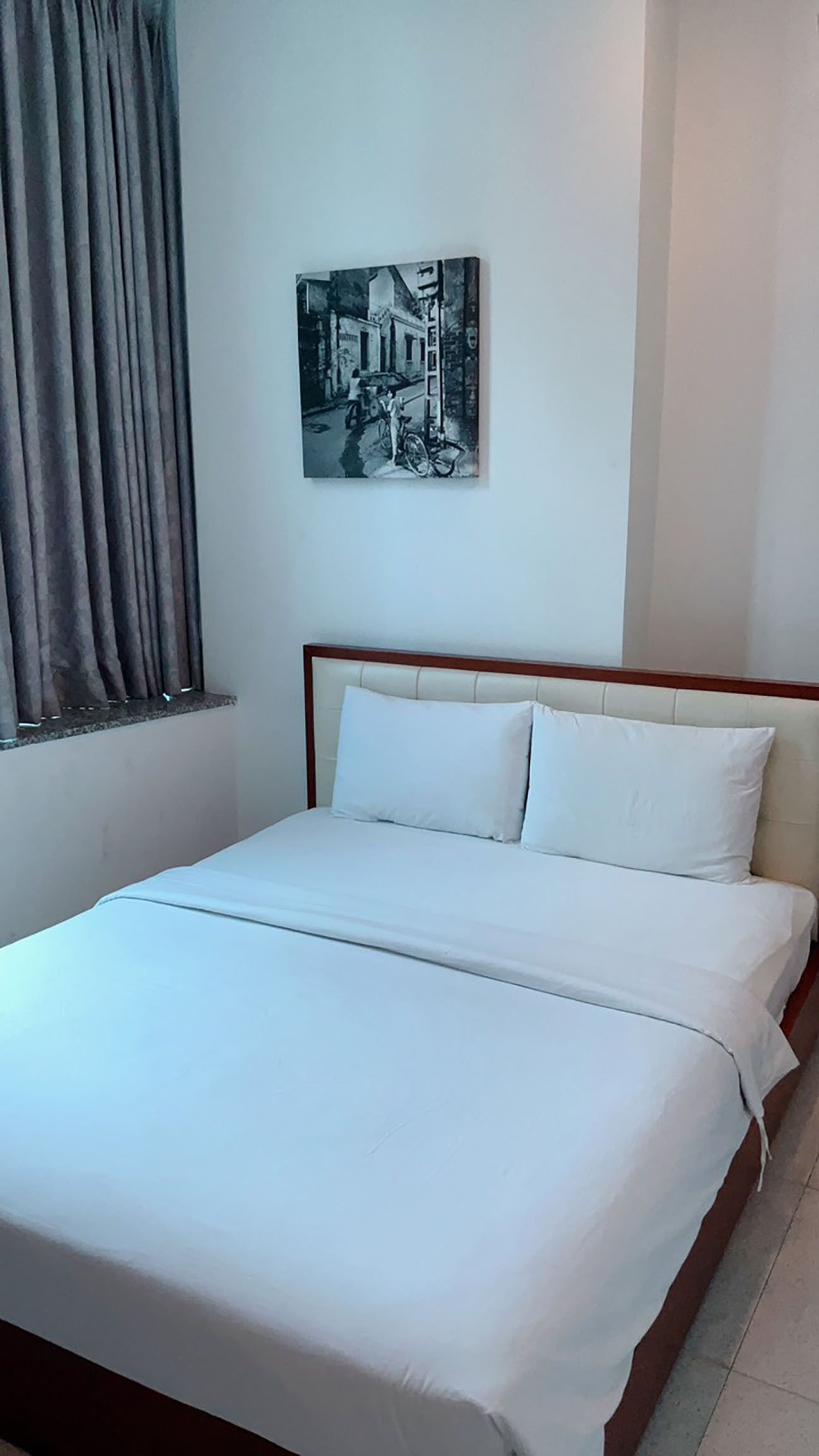 Un dormitorio con cama, ropa de cama blanca, cabecero, ventana con cortinas y un cuadro colgado en la pared.