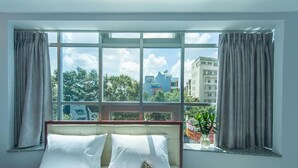 A hotel room with a large window, a bed with white linens, and a view of buildings and trees outside. - Nu hotel (Ho Chi Minh City)