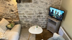 A cozy living room with a stone wall, a white sofa, a grey armchair, a small white table, and a television on a cabinet.