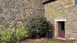 A stone building with a red door, a small garden with red flowers, and a metal chair outside.