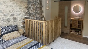 A bedroom with a stone wall, a wooden bed, a nightstand, a round mirror with a light, and a wooden wardrobe.