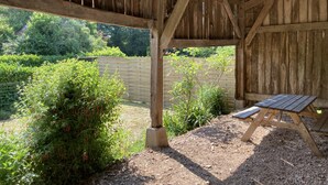 A wooden pergola with a bench, surrounded by dense greenery and a wooden fence.