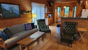 L’intérieur d’un chalet confortable, avec des murs en rondins de bois, un canapé, des fauteuils, un coin repas et une tête de cerf accrochée au mur.
