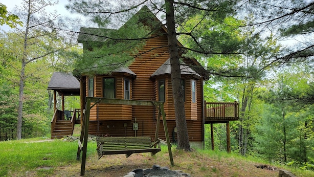 Une cabane rustique en rondins de bois, dotée d’un porche, d’une balançoire et d’un foyer extérieur.