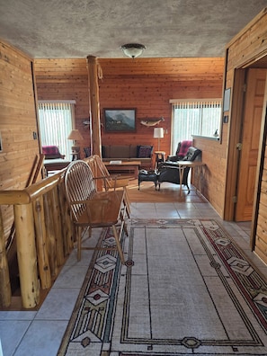 A log cabin interior with a sofa, chairs, a dining table, a rug, and a mounted deer head.