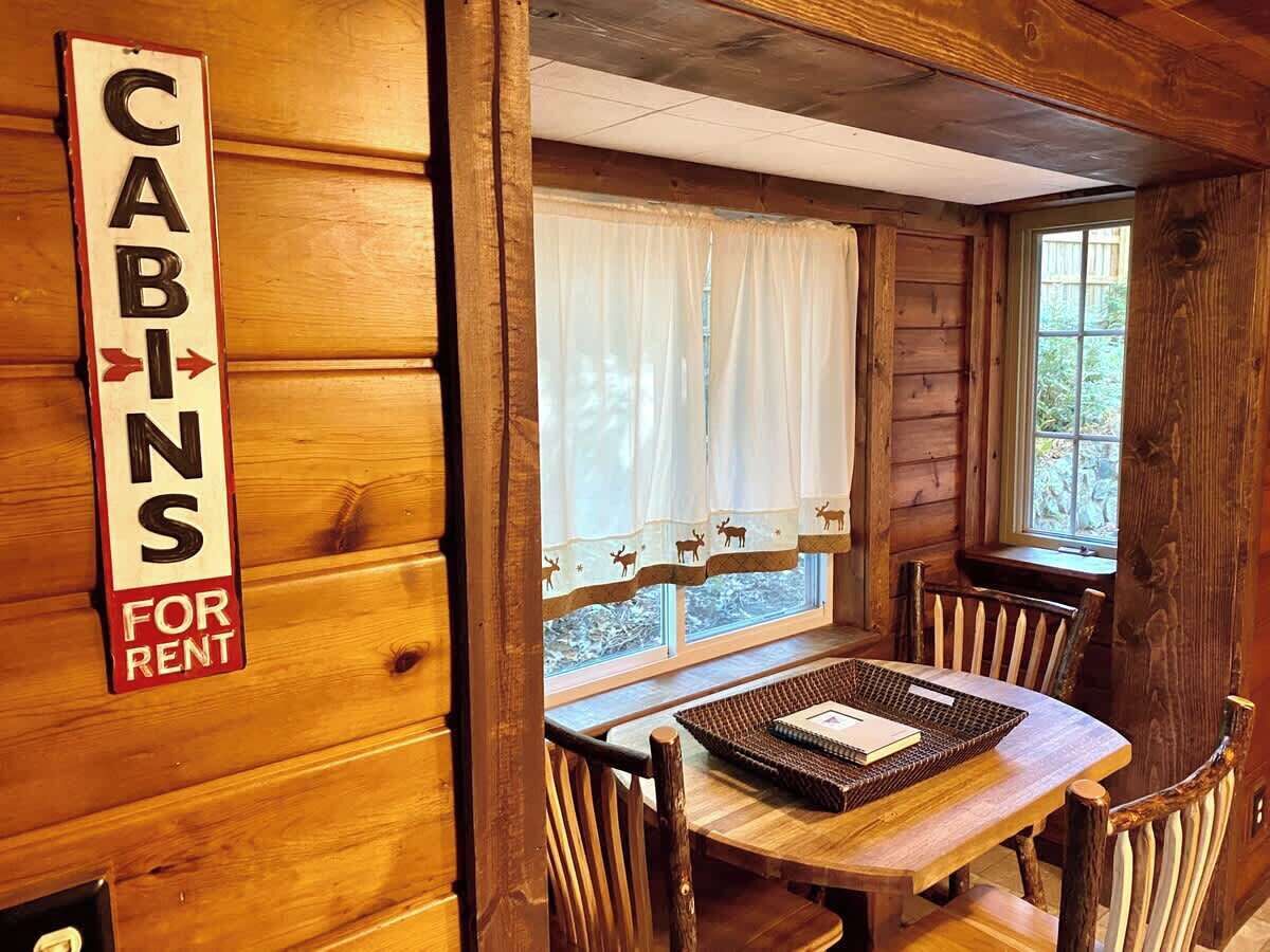 A cabin interior with a table and chairs, a window with curtains, and a sign indicating cabins for rent.