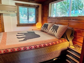 Una habitación acogedora estilo cabaña con una cama, una ventana con persianas, una lámpara y una mesita de noche de madera.