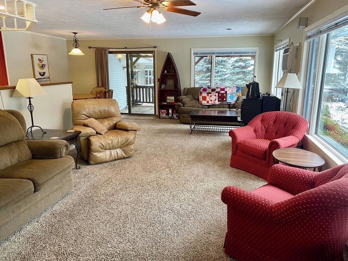 A cozy living room with a beige leather sofa, a red armchair, and a coffee table. A bookshelf and a window with a view of trees are also visible.