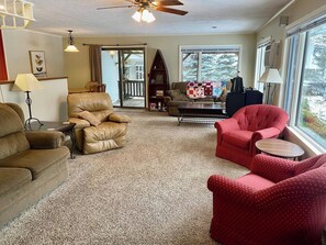 A cozy living room with a beige leather sofa, a red armchair, and a coffee table. A bookshelf and a window with a view of trees are also visible. - Secluded Riverfront Home in Grayling (Grayling)