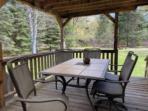 A covered outdoor patio with a table and chairs, surrounded by a wooden deck and a view of trees.