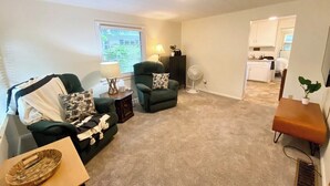 A living room with a sofa, armchair, and a small table with a lamp. There is a window with blinds, a fan, and a kitchen area in the background. - Convenient 2 Bedroom Home in Traverse City (Traverse City)
