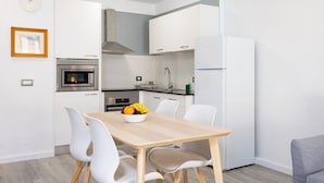 A modern kitchen with white cabinets, a stainless steel oven, a built-in microwave, and a refrigerator. There is a wooden dining table with white chairs and a bowl of fruit on it.