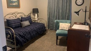 A bedroom with a metal bed frame, a blue and white checkered bedspread, a teal armchair, a small wooden desk, a lamp, a clock, and a framed picture on the wall.