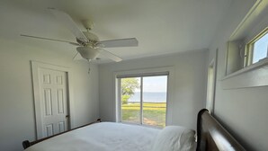 A bedroom with a bed, a ceiling fan, a window with a view of the sea, and a door.