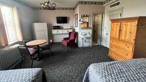 Una habitación de hotel con cama, escritorio con silla, kitchenette pequeño, televisor y armario.