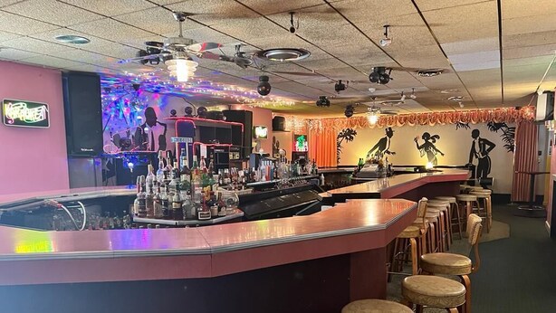 Una zona de bar con mostrador, taburetes y una variedad de botellas y vasos.