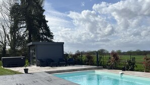 Une piscine avec une terrasse en bois, un petit abri de jardin et une zone herbeuse agrémentée d’une plante en pot.