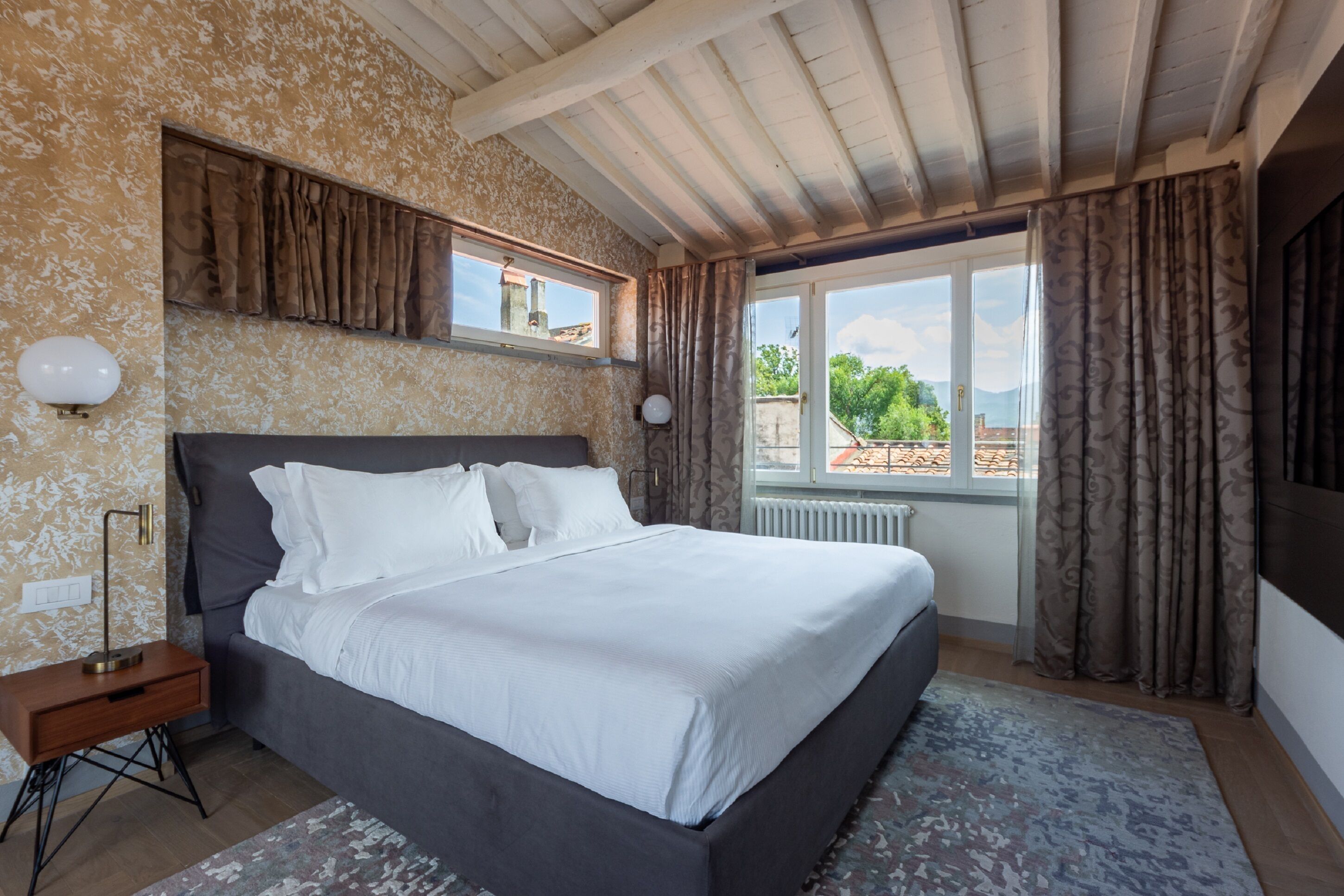 A bedroom with a large bed, a nightstand, a window with curtains, and a view of the outside.