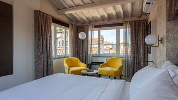 Quarto de hotel com uma cama grande, duas poltronas amarelas, uma mesa pequena e vista para prédios através de amplas janelas.