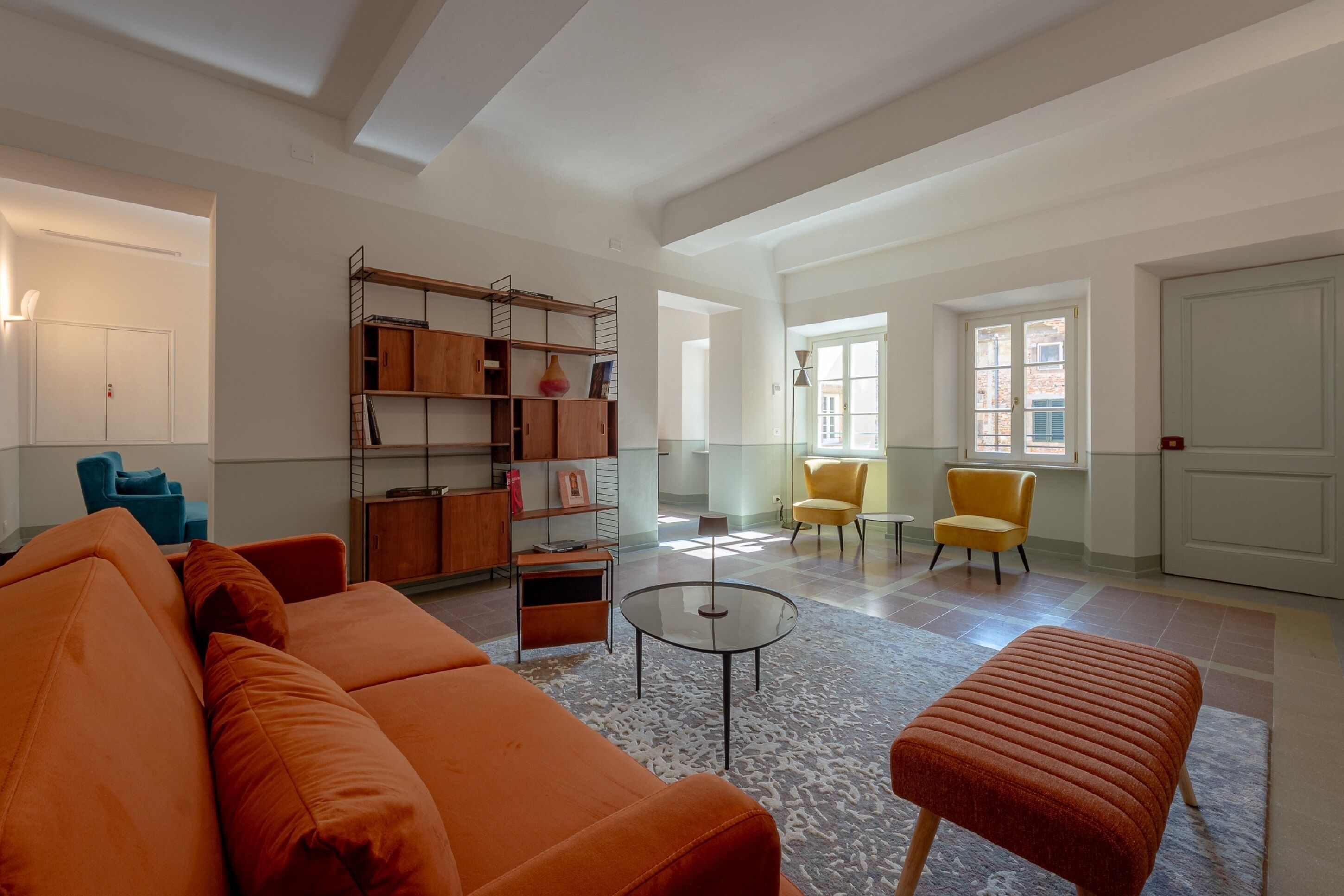 Un vaste salon avec un grand canapé orange, une table basse en verre et une bibliothèque. On y trouve deux fauteuils jaunes et un fauteuil bleu. La pièce est dotée d’un haut plafond avec des poutres apparentes et de grandes fenêtres.