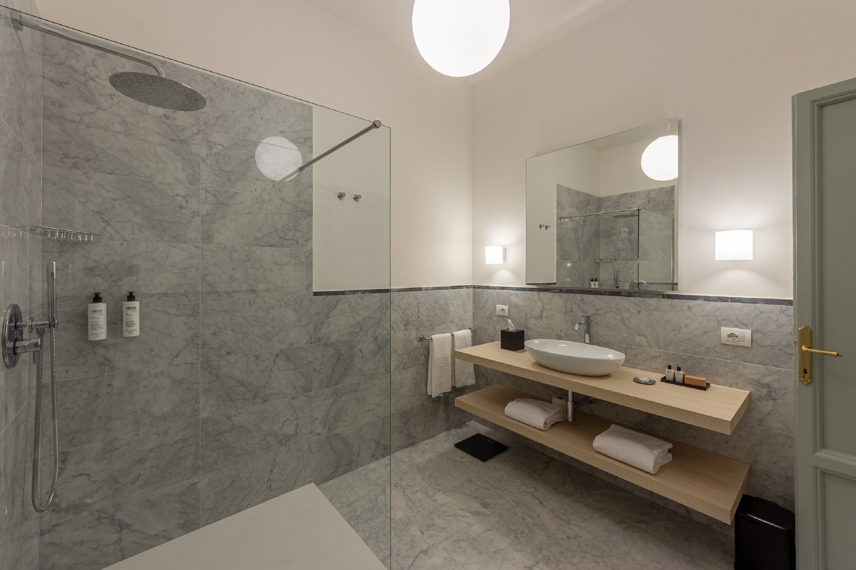 Un baño moderno con ducha de vidrio, pared con azulejos de mármol, lavamanos blanco y tocador de madera.