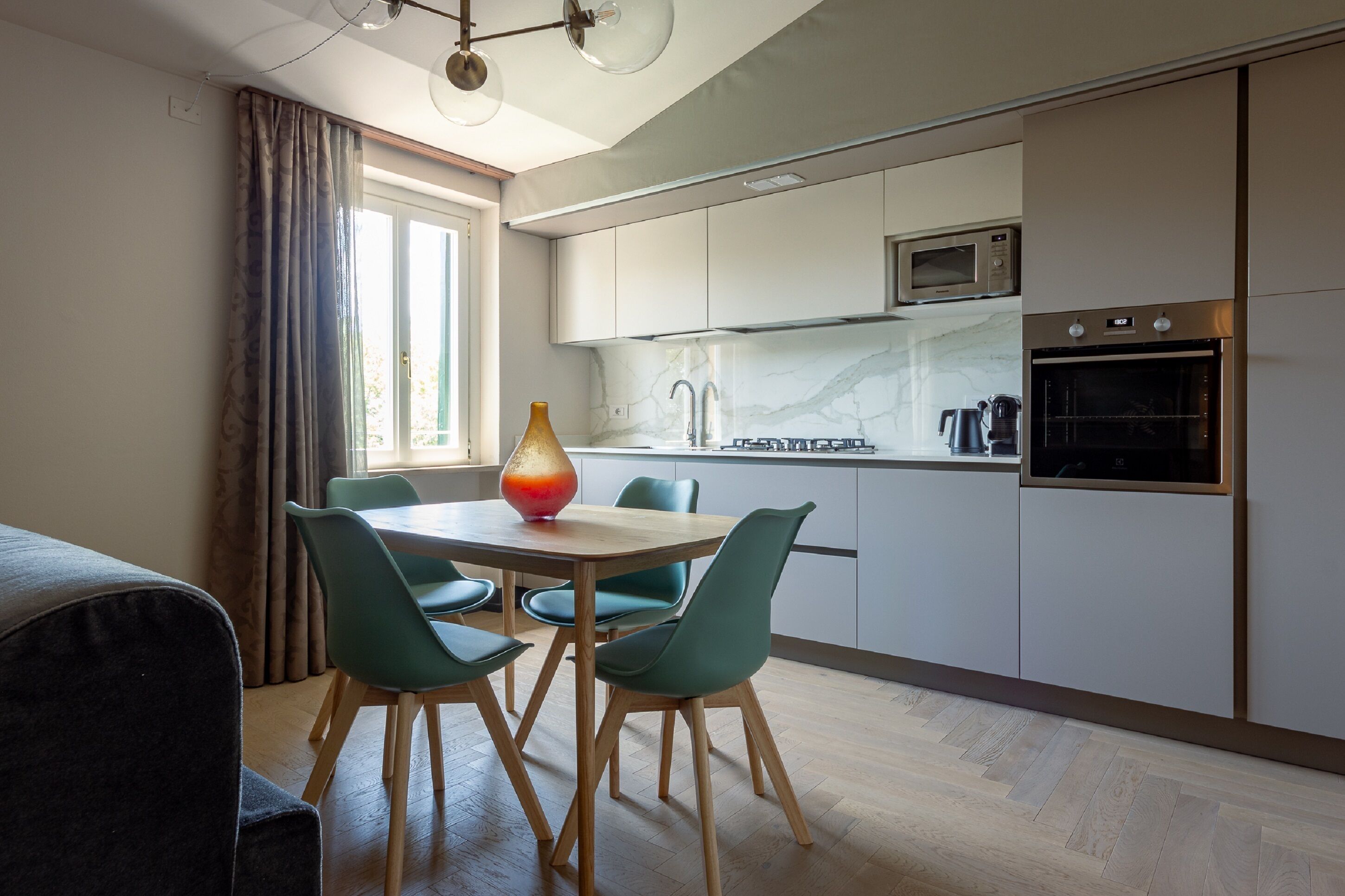 A modern kitchen with white cabinets, a microwave, and an oven. There is a dining table with chairs and a vase on it.