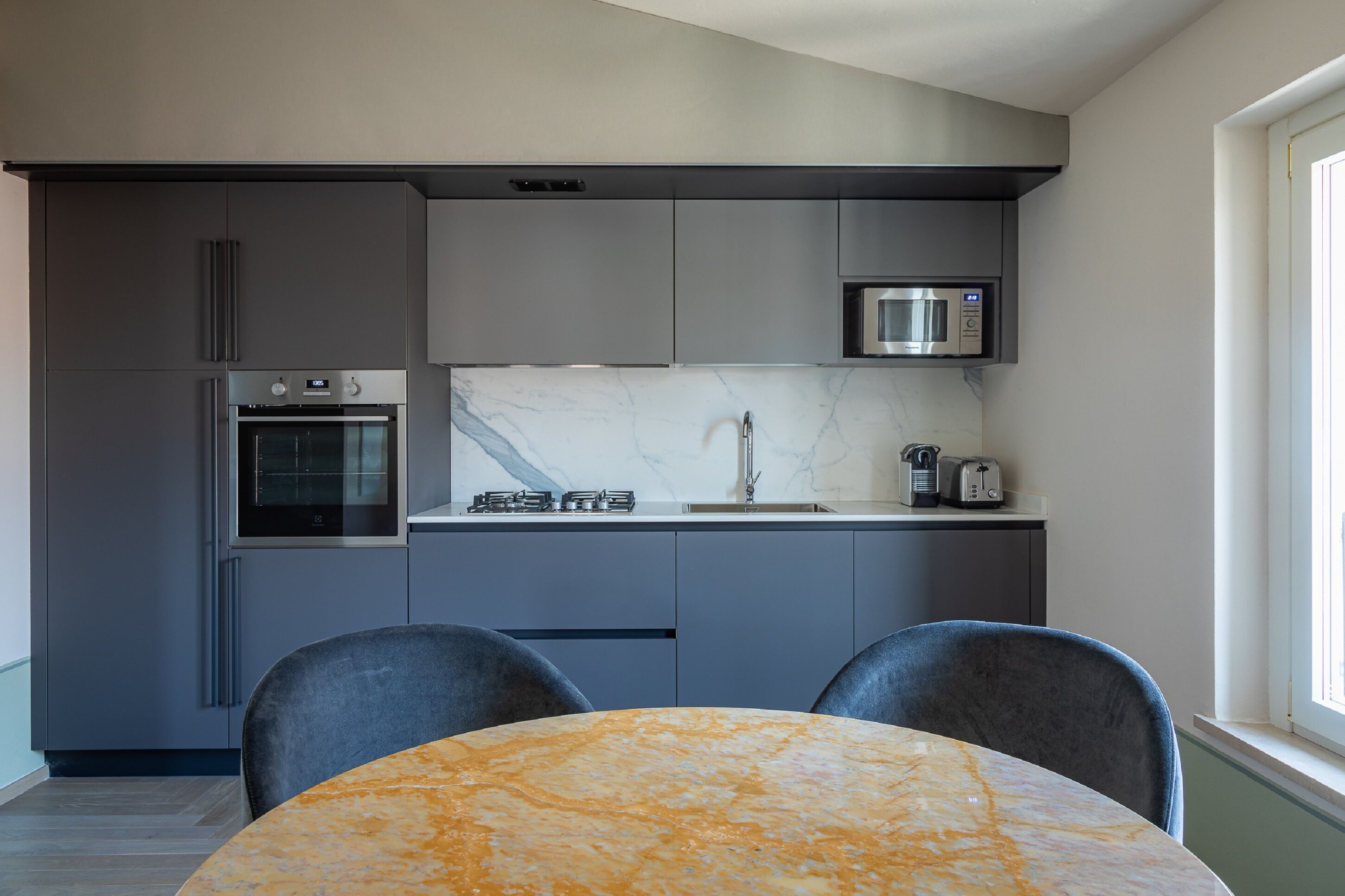 Une cuisine moderne avec des meubles gris, un four encastré, un micro-ondes et un pare-feu en marbre. On aperçoit au premier plan une table ronde avec deux chaises.