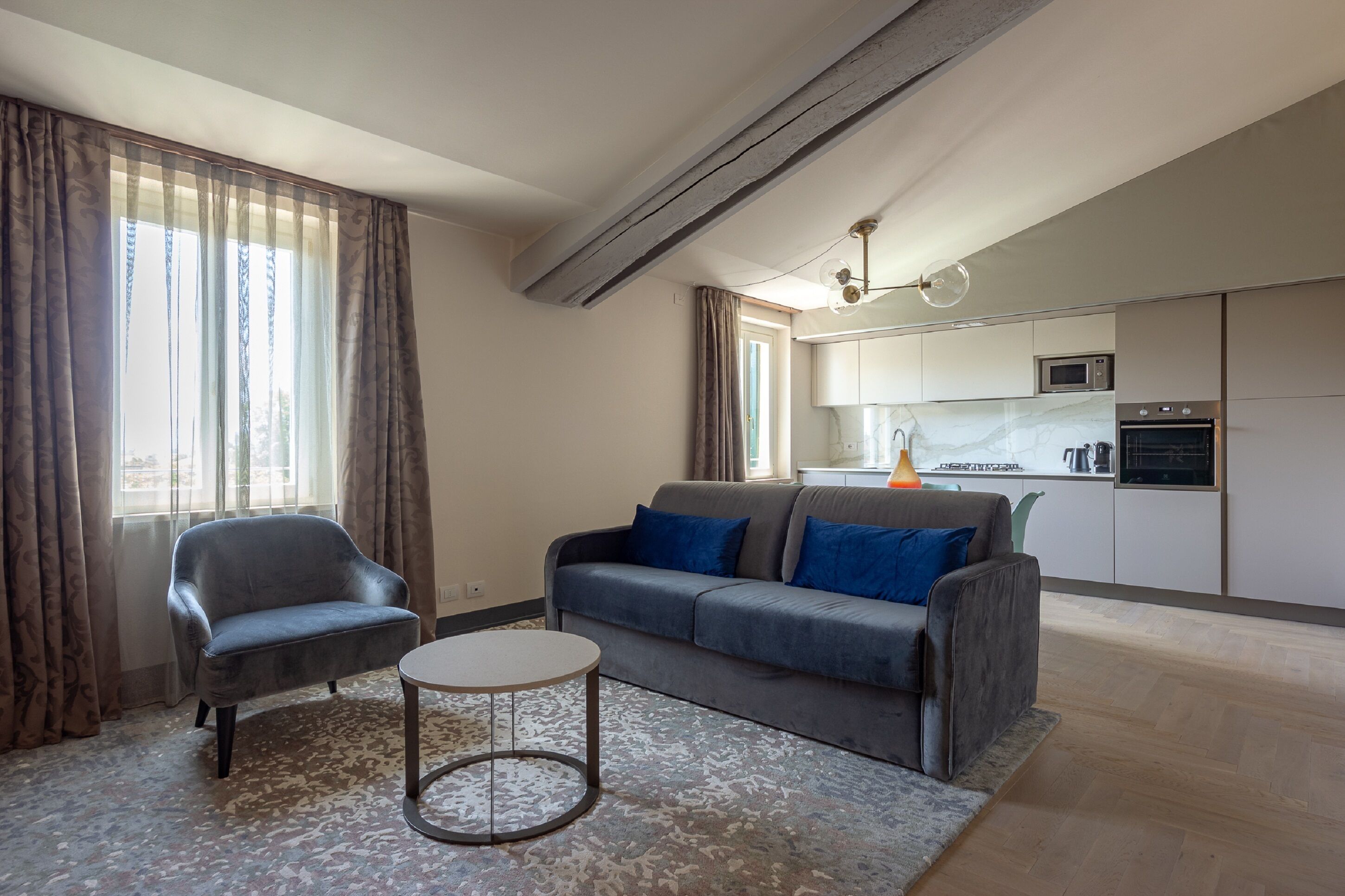 Un soggiorno moderno con un divano grigio, un tavolino rotondo e una poltrona. Si intravede in secondo piano una cucina con forno a microonde e forno.