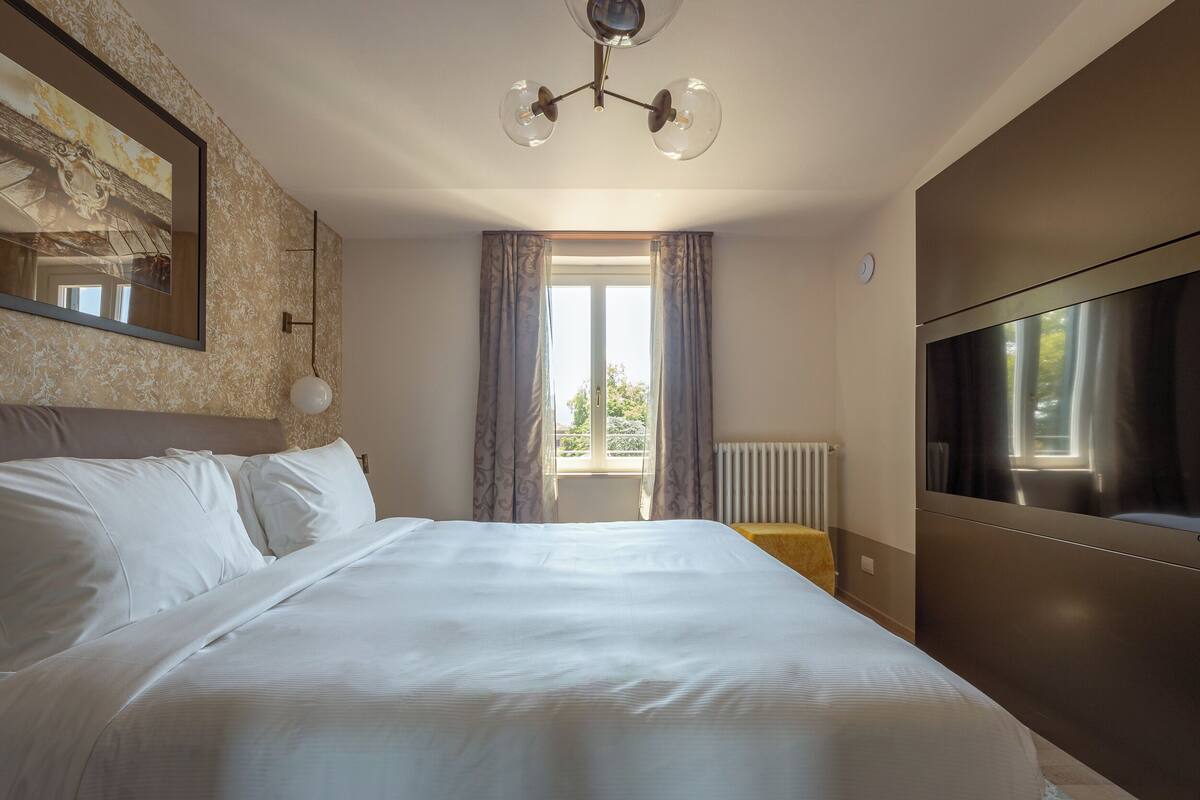 Ein Schlafzimmer mit einem großen Bett, einer Deckenleuchte, einem Fenster mit Vorhängen, einem Heizkörper und einem Fernseher an der Wand.