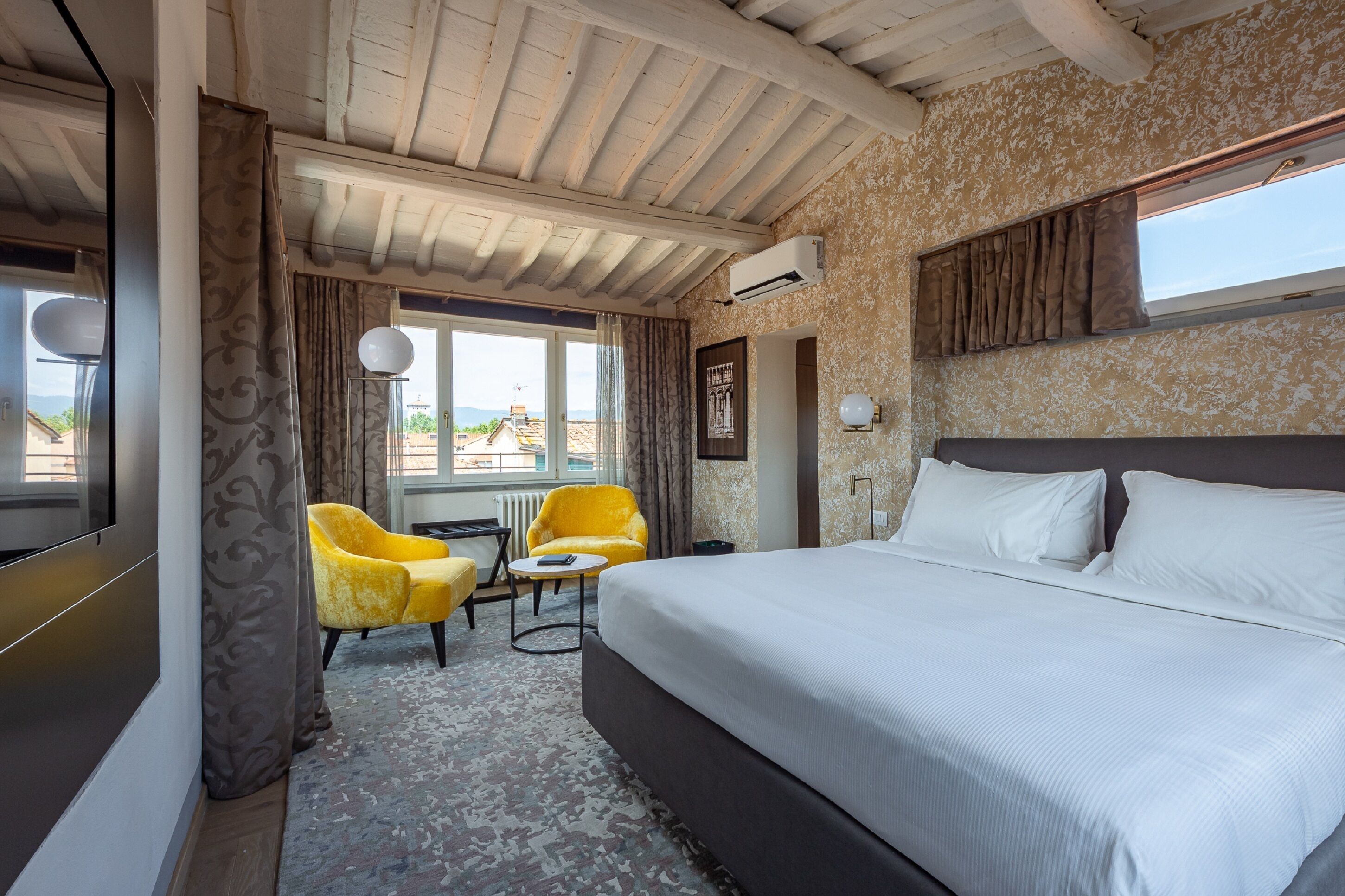 Camera d'albergo con un letto grande, un tavolino, due poltrone gialle, una finestra con tende e vista sull'esterno.