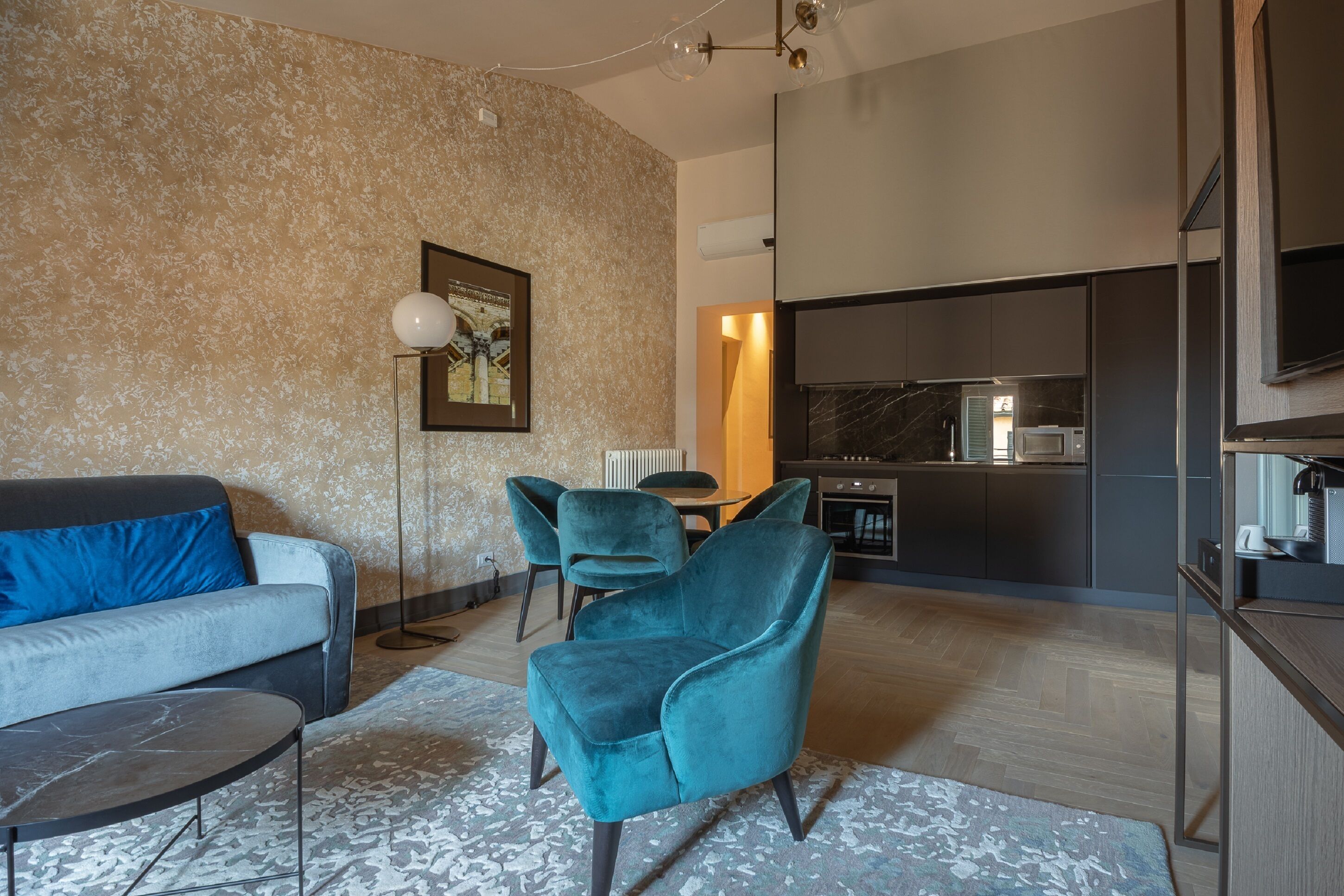 A modern living room with a sofa, teal armchairs, a coffee table, and a floor lamp. A kitchen is visible in the background.