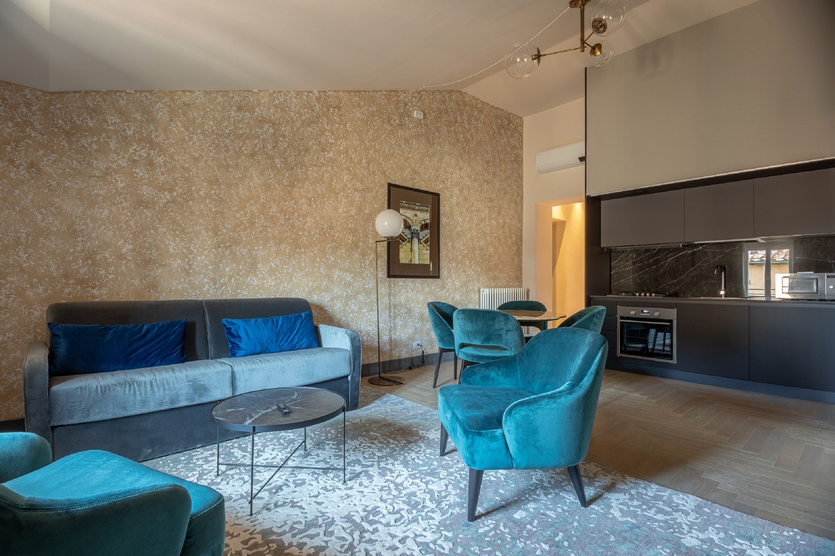 Una sala moderna con un sofá gris, sillones azul verdoso y una mesa de centro. Se ve una cocina al fondo con armarios oscuros y una encimera de mármol.