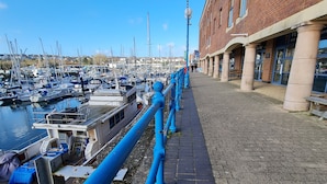 Una passeggiata lungomare con una fila di barche ormeggiate a sinistra, un edificio in mattoni con colonne a destra e un cielo azzurro sopra.