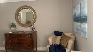 A room with a wooden dresser, a round mirror, a beige armchair with a blue throw blanket, and a white side table. - Bettendorf Condo: 2BR, 2BA, Garage, King master, Pets Welcome! (Bettendorf)
