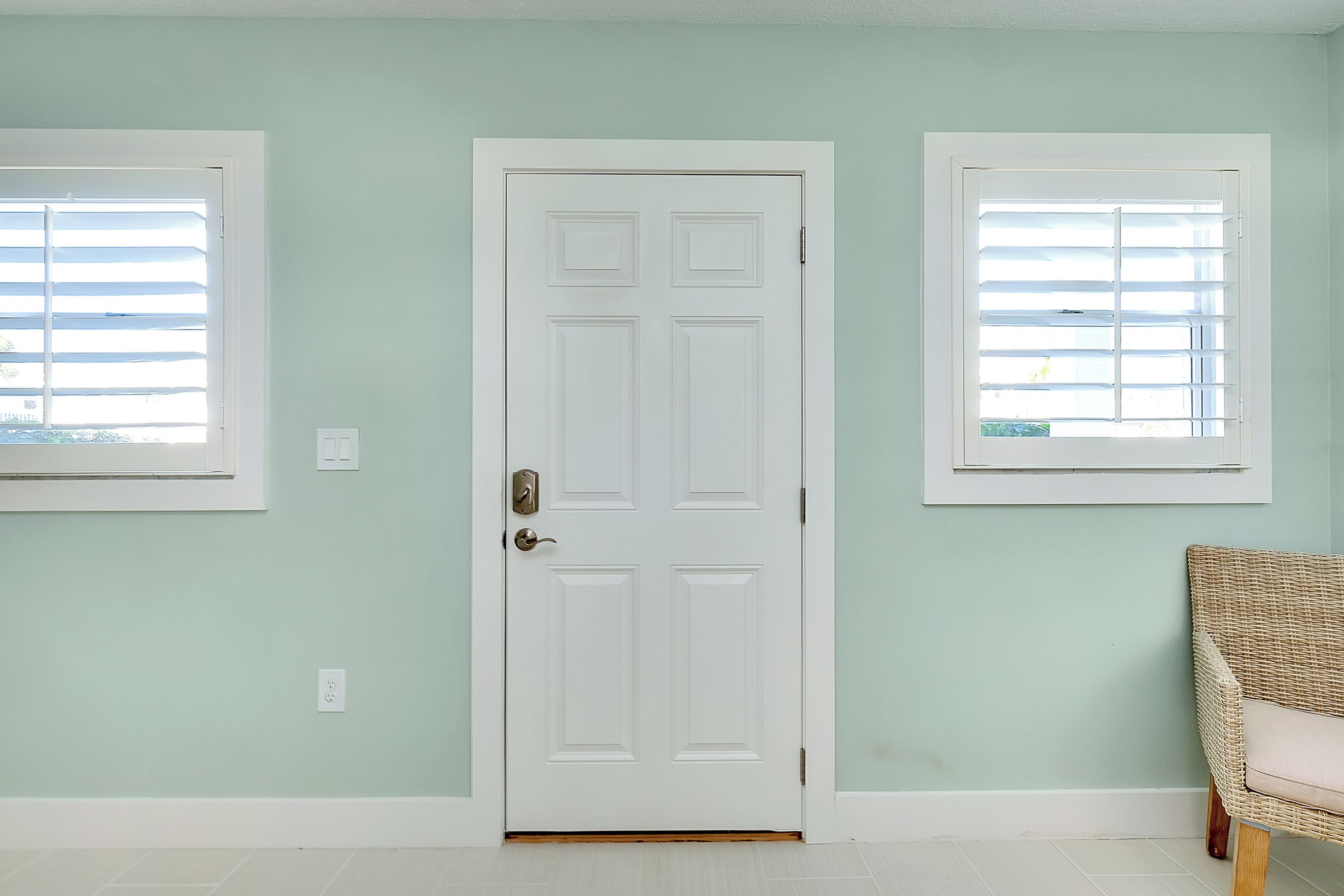 A room with a white door, two windows with blinds, and a wicker chair.