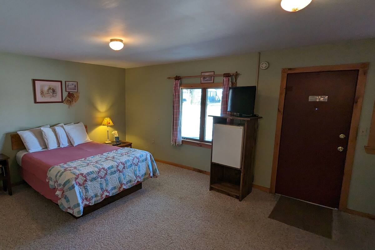 A bedroom with a bed, bedside lamps, a TV on a stand, a door, and a window with curtains.
