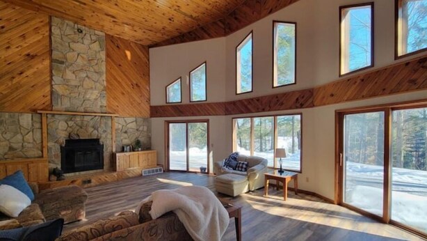 A spacious living room with a stone fireplace, wooden beams, and large windows offering views of a snowy landscape. - Waterfront Peninsula Escape Cabin (Lakewood)