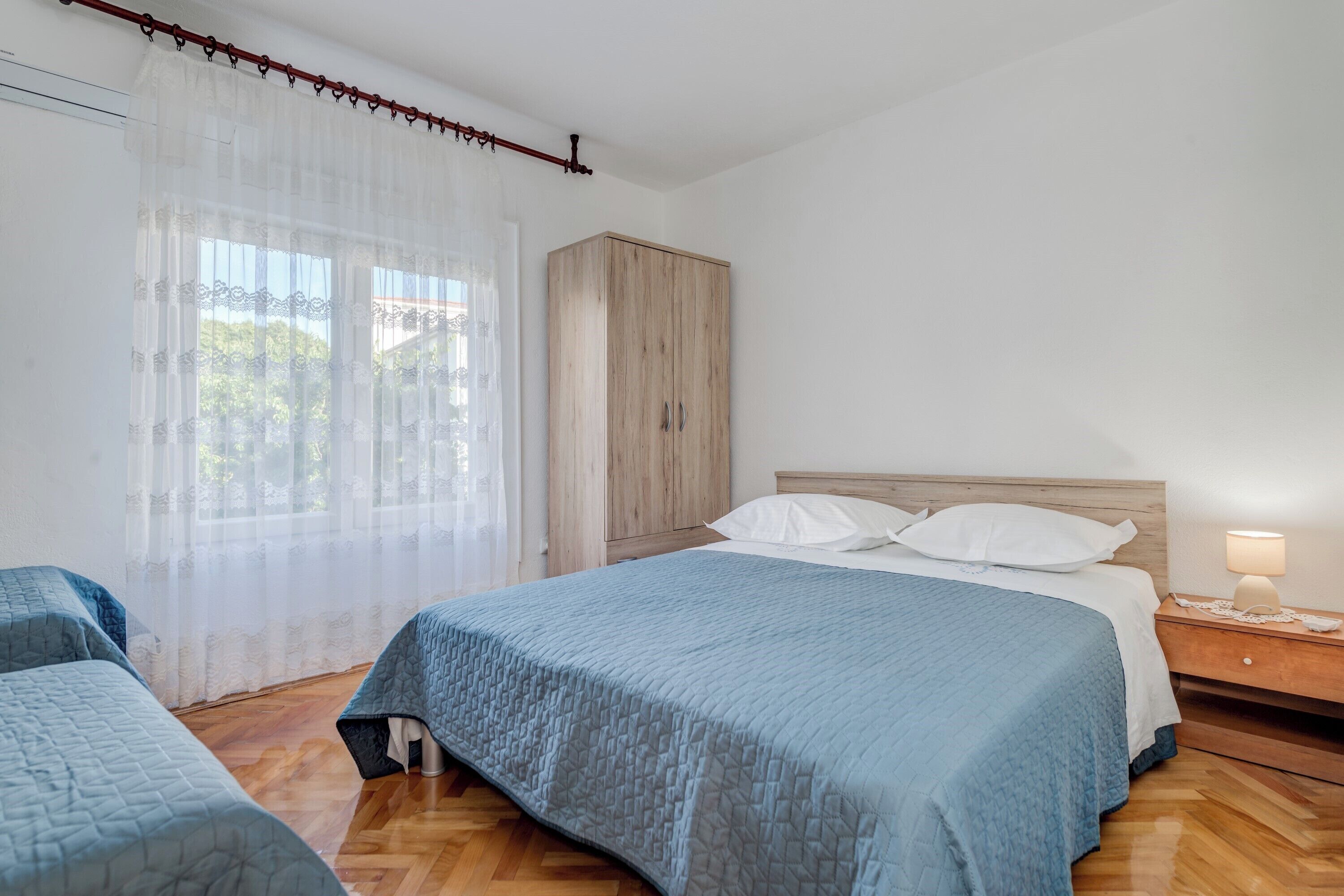 Ein Schlafzimmer mit einem Bett, zwei Nachttischen, einem Kleiderschrank und einem Fenster mit durchsichtigen Vorhängen.