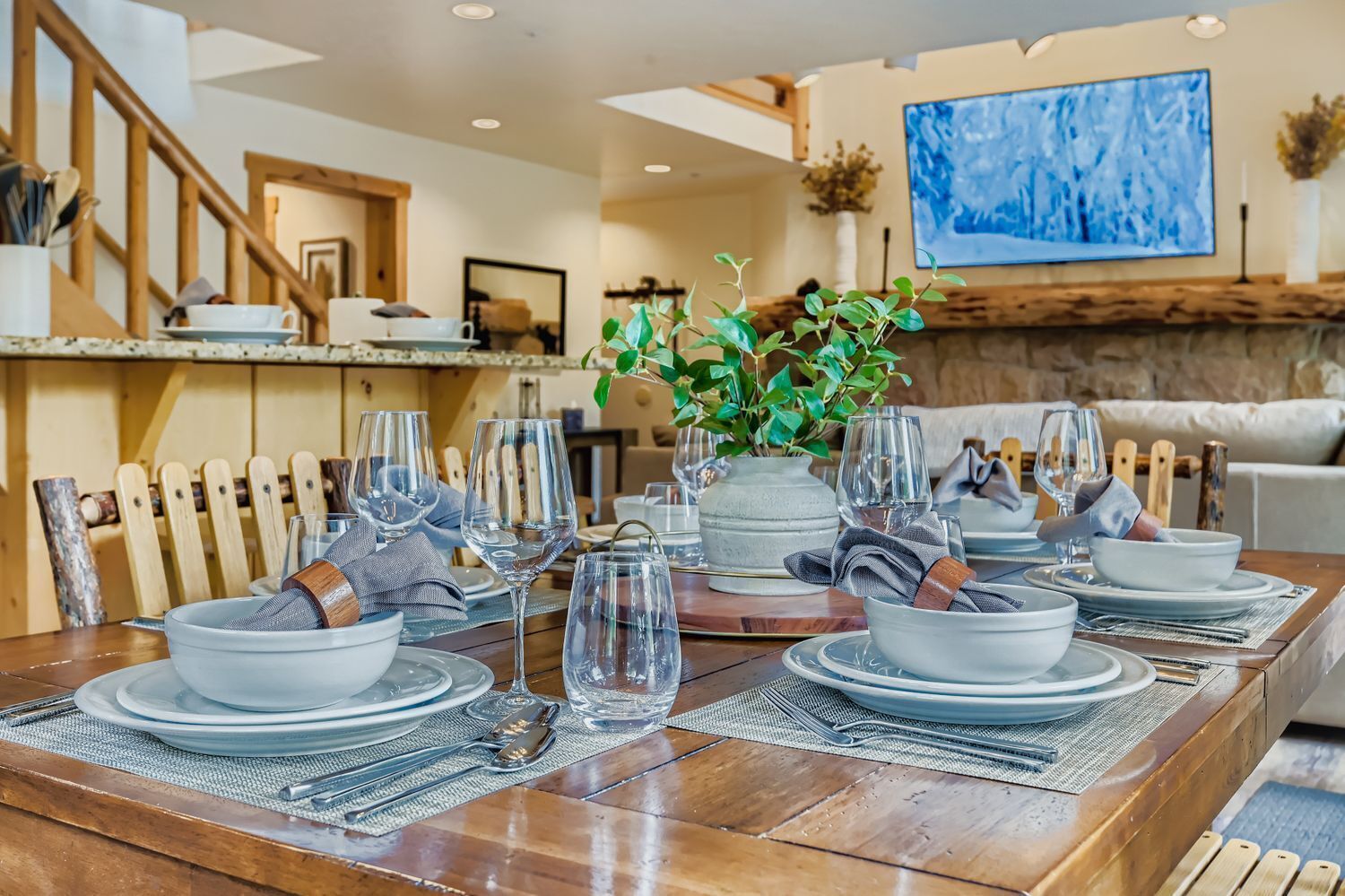 A dining table set with plates, glasses, and cutlery, with a plant centerpiece and a painting on the wall.