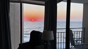 Una stanza con vista sul tramonto sull'oceano, un balcone con sedie e un tavolo con una lampada.