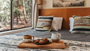 A cozy room with a bed, a window with a view of trees, a coffee table with a mug, a plate of pastries, and a wall-mounted basket.