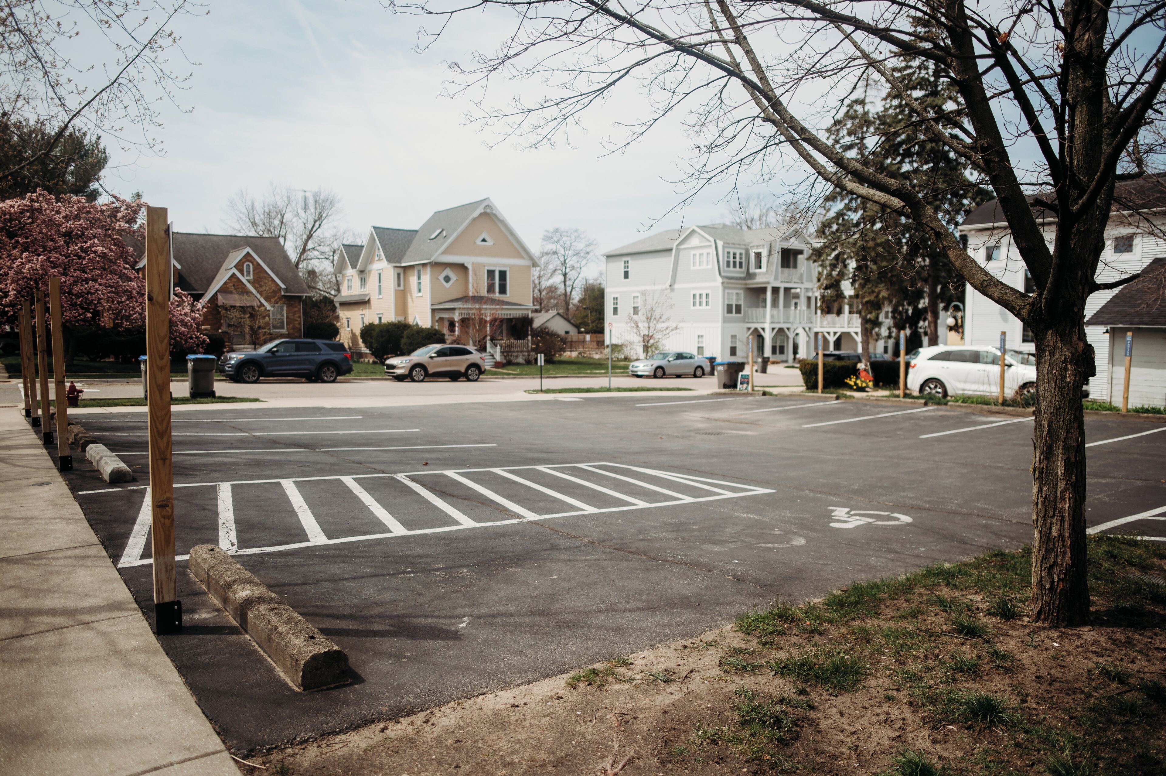 A parking lot with marked spaces and bollards, surrounded by residential houses and a tree.