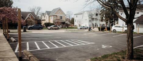 A parking lot with marked spaces and bollards, surrounded by residential houses and a tree.