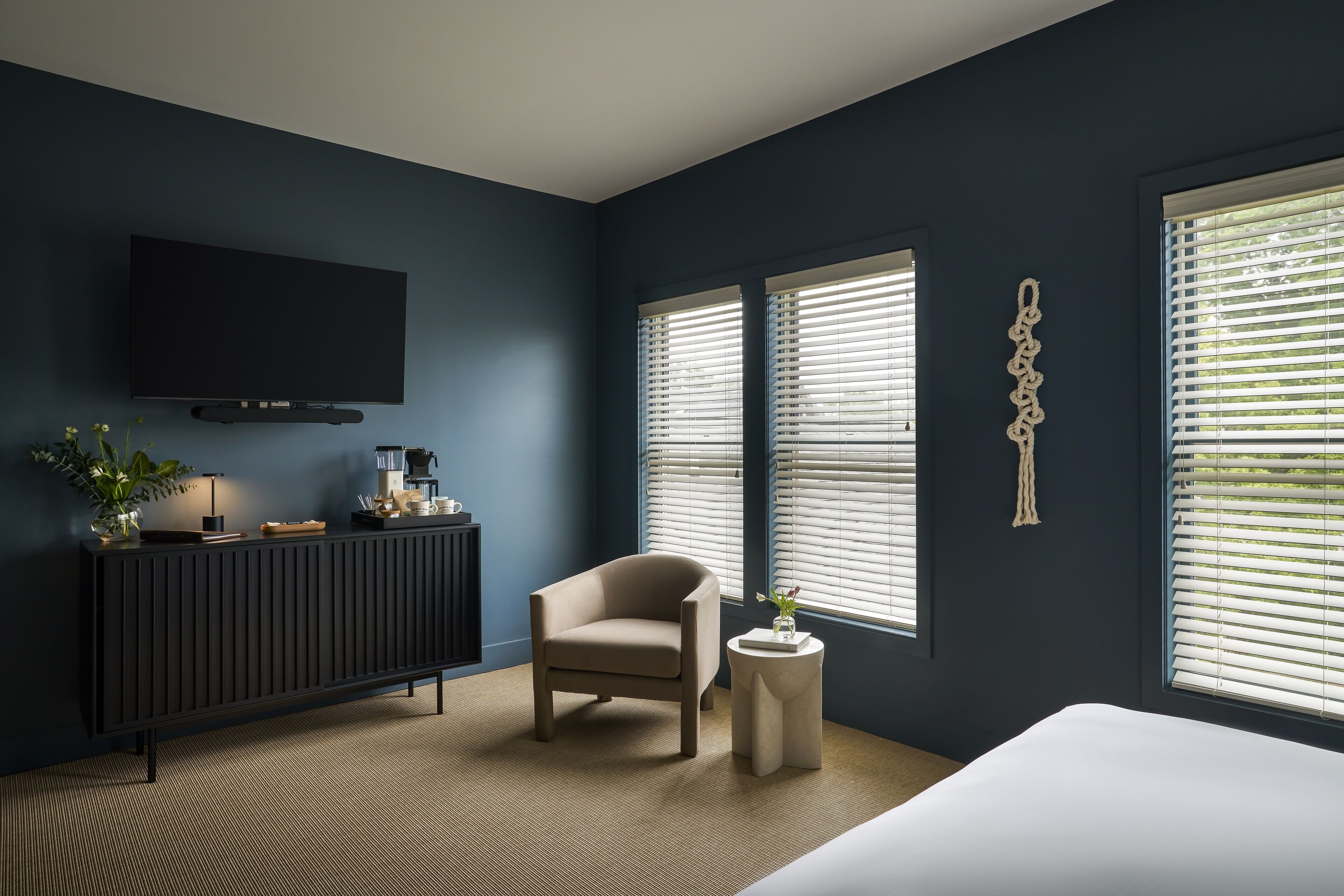 A modern bedroom with a dark blue wall, a large bed, a TV mounted on the wall, and a window with blinds.