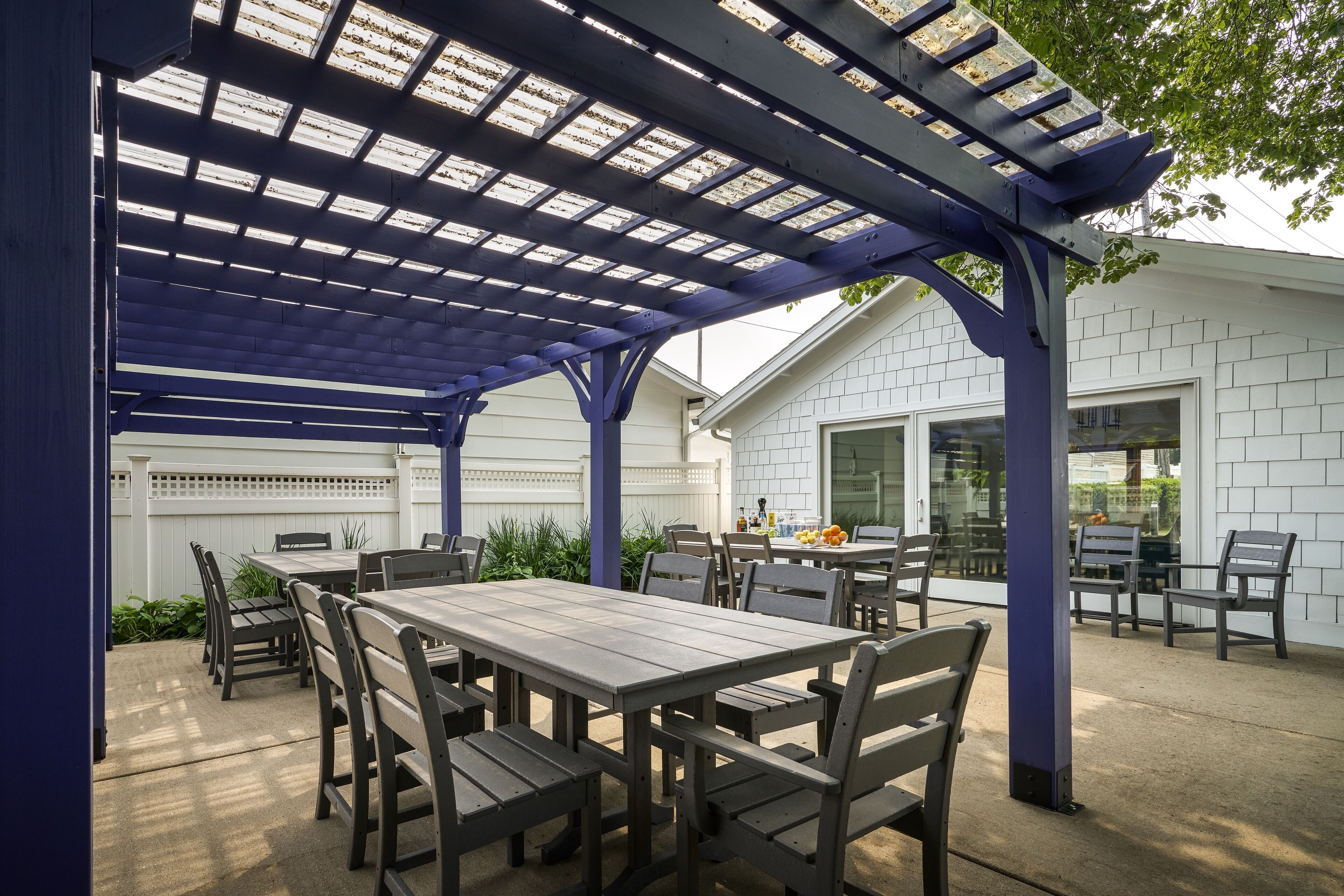 A covered patio area with a wooden table and chairs, a pergola with blue beams, and a house in the background.