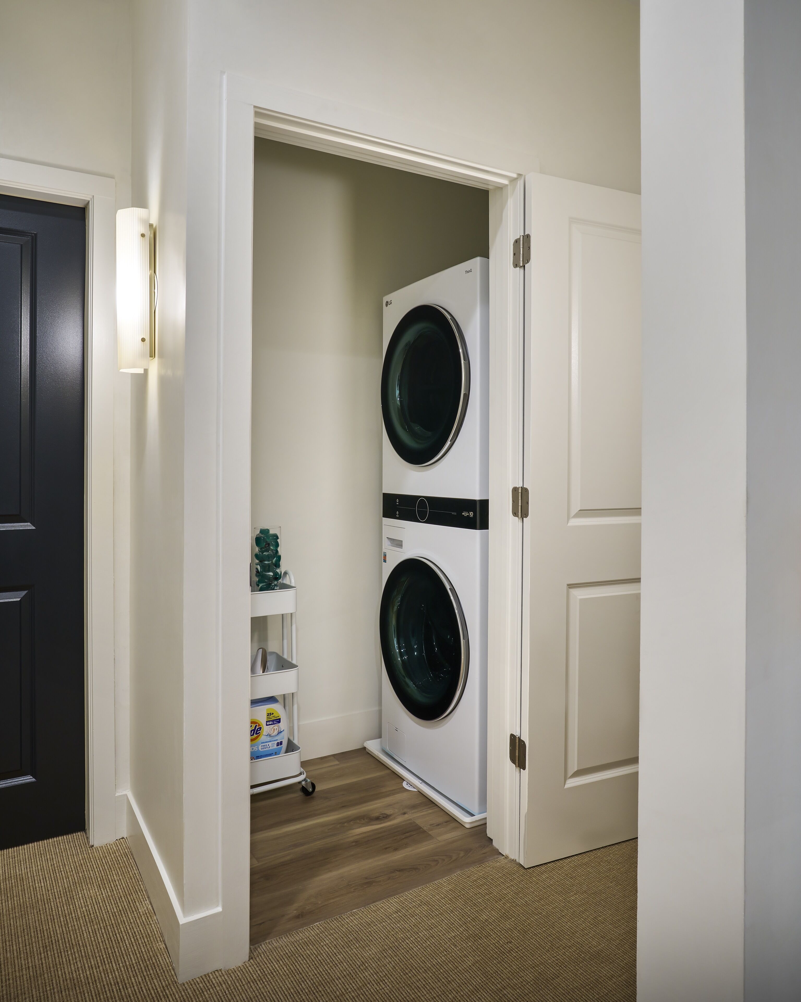 A laundry room with two stacked front-loading washing machines, a white shelving unit with laundry supplies, and a dark door.